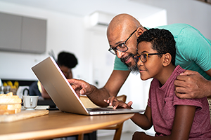 Man helping young boy with laptop computer at kitchen table